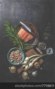 Raw salmon fillet on wooden plate with dill, knife and other ingredients for preparing a delicious fish dish: onions, shrimp on cutting board and jars of dry seasoning. View from above