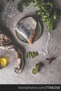 Raw salmon steak with sheds at plate on grey concrete kitchen table with salt, parsley, lime, olive oil and kitchen cloth. Healthy cooking at home with fresh fish. Top view.