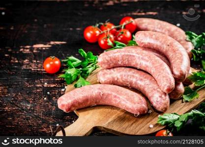 Raw sausages on a wooden cutting board with tomatoes and herbs. Against a dark background. High quality photo. Raw sausages on a wooden cutting board with tomatoes and herbs.