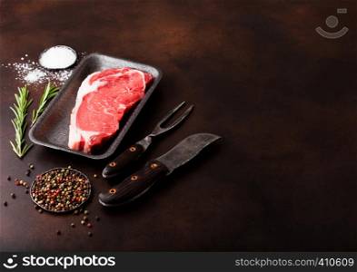 Raw sirloin beef steak in plastic tray with knife and fork on rusty background. Salt and pepper in round plates
