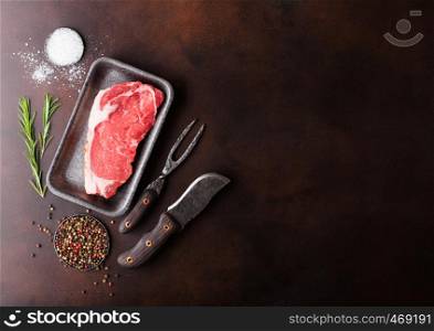 Raw sirloin beef steak in plastic tray with knife and fork on rusty background. Salt and pepper in round plates