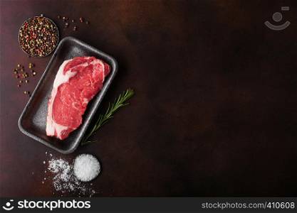 Raw sirloin beef steak in plastic tray with salt and pepper and fresh rosemary on rusty board
