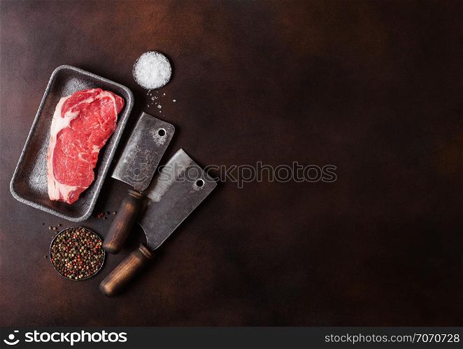 Raw sirloin beef steak in plastic tray with salt and pepper and vintage meat hatchets on rusty board.