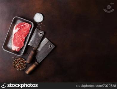 Raw sirloin beef steak in plastic tray with salt and pepper and vintage meat hatchets on rusty board.