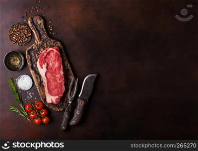 Raw sirloin beef steak on old vintage chopping board with knife and fork on rusty background. Salt and pepper with fresh rosemary and tomatoes.