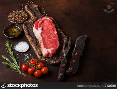 Raw sirloin beef steak on old vintage chopping board with knife and fork on rusty background. Salt and pepper with fresh rosemary and tomatoes.