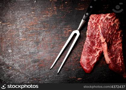 Raw steak with a large fork on the table. Against a dark background. High quality photo. Raw steak with a large fork on the table.