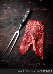 Raw steak with a large fork on the table. Against a dark background. High quality photo. Raw steak with a large fork on the table.