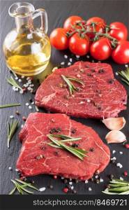 Raw steak with rosemary, salt and pepper on black slate background. ingredients for cooking beefsteak, tomato, olive oil, rosemary, spices. Vertical orientation. Fresh raw beef steak with spice on black background