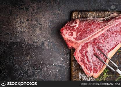 Raw T-bone Steak for grill or BBQ with meat fork on aged cutting board and dark rustic metal background, top view