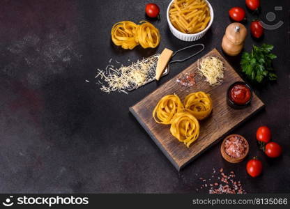 Raw tagliatelle paste with grated cheese, cherry tomatoes, spices and herbs on a wooden cutting board against a dark concrete background. Raw tagliatelle paste with grated cheese, cherry tomatoes, spices and herbs on a wooden cutting board