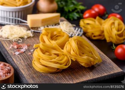 Raw tagliatelle paste with grated cheese, cherry tomatoes, spices and herbs on a wooden cutting board against a dark concrete background. Raw tagliatelle paste with grated cheese, cherry tomatoes, spices and herbs on a wooden cutting board