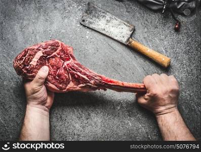 Raw tomahawk beef steak in male hands with meat cleaver on dark rustic background. Butcher hands holding tomahawk steak, top view