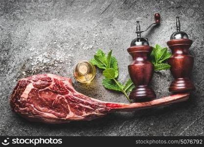 Raw tomahawk beef steak with condiment , salt and pepper shakers or mill and fresh herbs on dark rustic concrete background, top view, place for text. Meat steak preparation