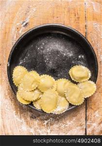 Raw Tortellini in black plate on wooden background with wheat flour, top view