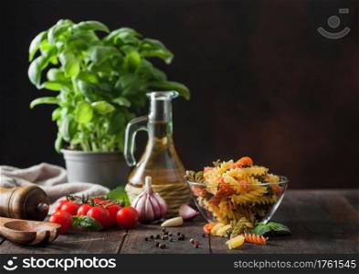 Raw tricolor fusilli pasta in glass bowl with oil and garlic, basil plant and tomatoes on wooden background. Space for text