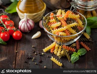 Raw tricolor fusilli pasta in glass bowl with oil and garlic, basil plant and tomatoes with pepper and linen towel on wooden table background.