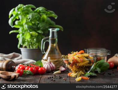 Raw tricolor fusilli pasta in glass bowl with oil and garlic, basil plant and tomatoes on wooden background. Space for text