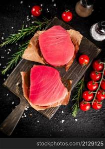 Raw tuna on a cutting board with tomatoes and rosemary. On a black background. High quality photo. Raw tuna on a cutting board with tomatoes and rosemary.
