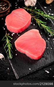 Raw tuna steak on a stone board with rosemary, spices and tomatoes. On a black background. High quality photo. Raw tuna steak on a stone board with rosemary, spices and tomatoes.