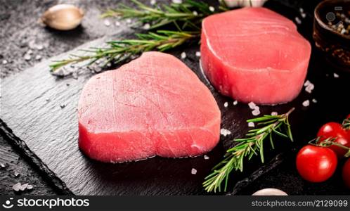 Raw tuna steak on a stone board with rosemary, spices and tomatoes. On a black background. High quality photo. Raw tuna steak on a stone board with rosemary, spices and tomatoes.