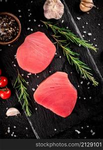 Raw tuna steak on a stone board with rosemary, spices and tomatoes. On a black background. High quality photo. Raw tuna steak on a stone board with rosemary, spices and tomatoes.