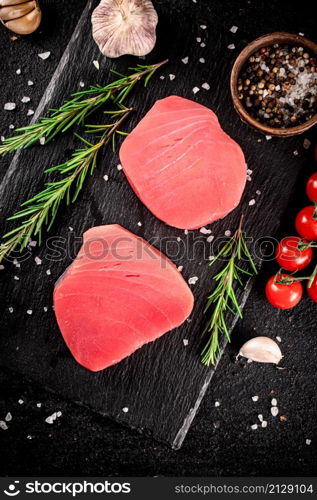 Raw tuna steak on a stone board with rosemary, spices and tomatoes. On a black background. High quality photo. Raw tuna steak on a stone board with rosemary, spices and tomatoes.