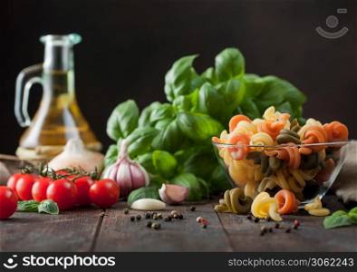 Raw wheat trottole pasta in glass bowl with basil plant, oil and tomatoes with garlic and pepper on wooden table background.