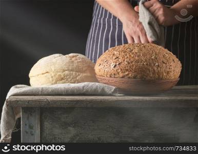 ray of light falls on baked round bread on a table and a woman in a striped apron, black background, concept of cooking