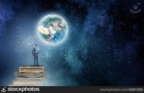 Reaching top of the world. Young confident businessman with arms on waist standing on pile of old books. Elements of this image are furnished by NASA