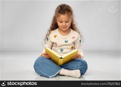 reading, education and childhood concept - beautiful smiling girl sitting on floor and reading book over grey background. beautiful smiling girl reading book on floor