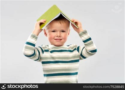 reading, education and childhood concept - portrait of smiling little boy with book on head as house roof top over grey background. portrait of smiling boy with book on head