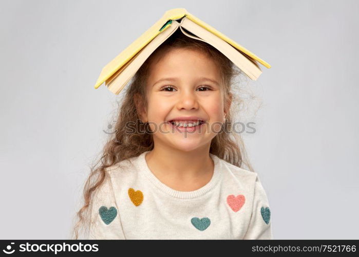 reading, education and childhood concept - portrait of smiling little girl with book on head as house roof top over grey background. portrait of smiling girl with book on head