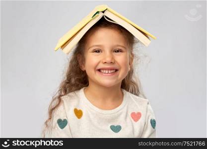 reading, education and childhood concept - portrait of smiling little girl with book on head as house roof top over grey background. portrait of smiling girl with book on head