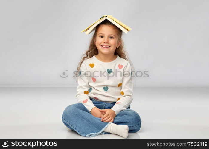 reading, education and childhood concept - portrait of smiling little girl with book on head as house roof top over grey background. portrait of smiling girl with book on head