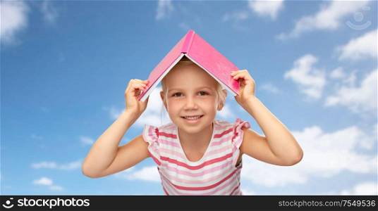 reading, education and childhood concept - smiling little girl with roof of book on top of her head over blue sky and clouds background. little girl with roof of book on top of her head