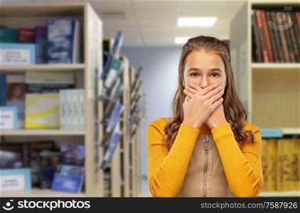reading, education and people concept - scared teenage student girl closing her mouth by hands over library background. scared student girl closing mouth at library