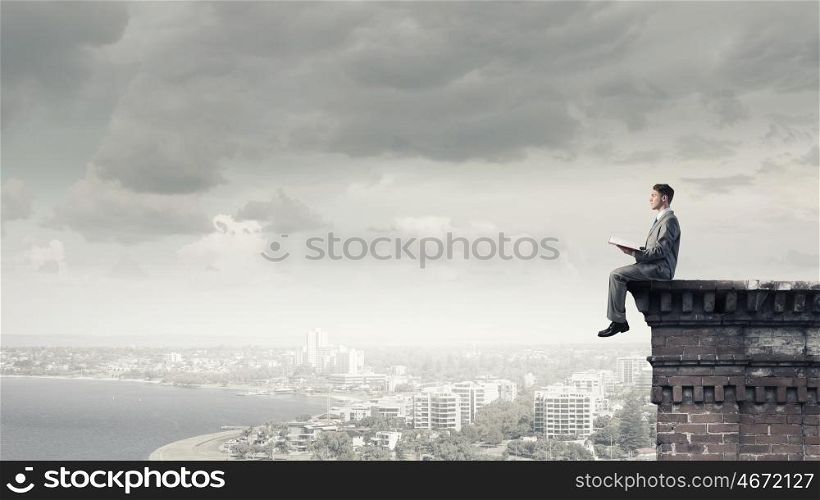 Reading in isolation. Young businessman sitting on building top and reading book