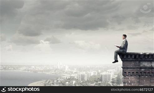Reading in isolation. Young businessman sitting on building top and reading book