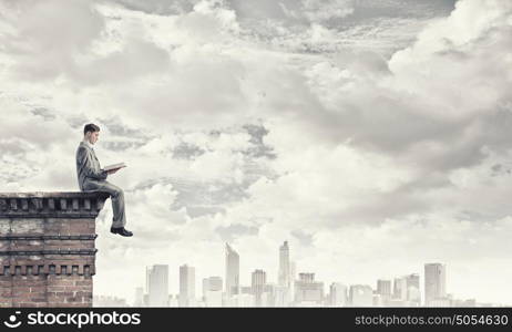 Reading in isolation. Young businessman sitting on building top and reading book