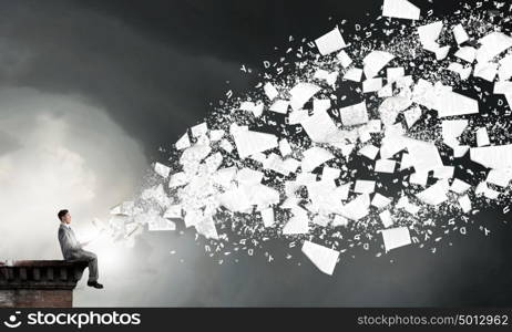 Reading in isolation. Young businessman sitting on building top with book and flying pages