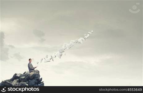 Reading in isolation. Young businessman sitting on rock top with opened book in hand and pages flying in air