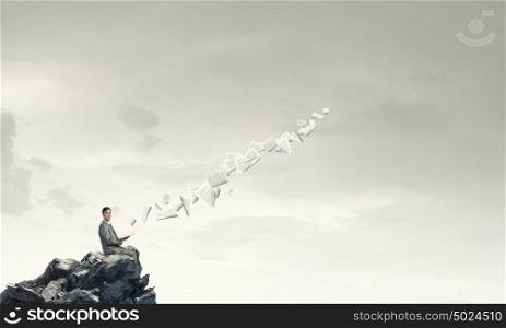 Reading in isolation. Young businessman sitting on rock top with opened book in hand and pages flying in air