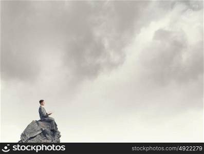 Reading in isolation. Young businessman sitting on rock top with opened book in hand