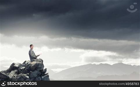 Reading in isolation. Young businessman sitting on rock top with opened book in hand