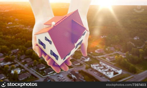 real estate, accommodation and property concept - close up of hands holding house or home model over suburbs background. close up of hands holding house or home model