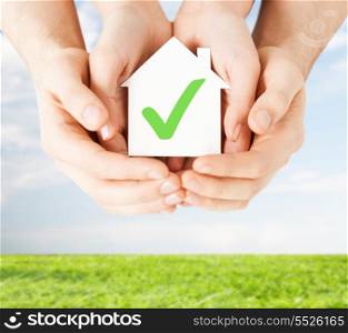 real estate and family home concept - male and female hands holding paper house with green check mark