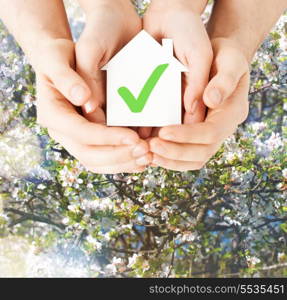 real estate and family home concept - male and female hands holding paper house with green check mark