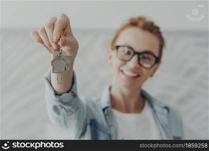 Real estate and relocation concept. Young happy smiling red-haired woman in casual clothes holding house keys of her new house home and showing it at camera, selective focus on female hand. Young happy smiling red-haired woman in casual clothes holding house keys of her new house home