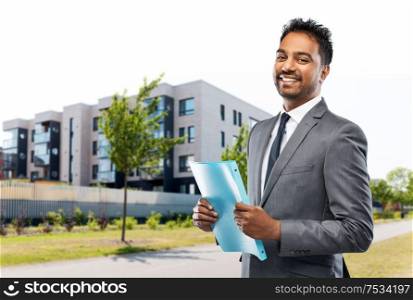 real estate business and people concept - indian man realtor with folder over living houses on city street background. indian man realtor with folder on city street
